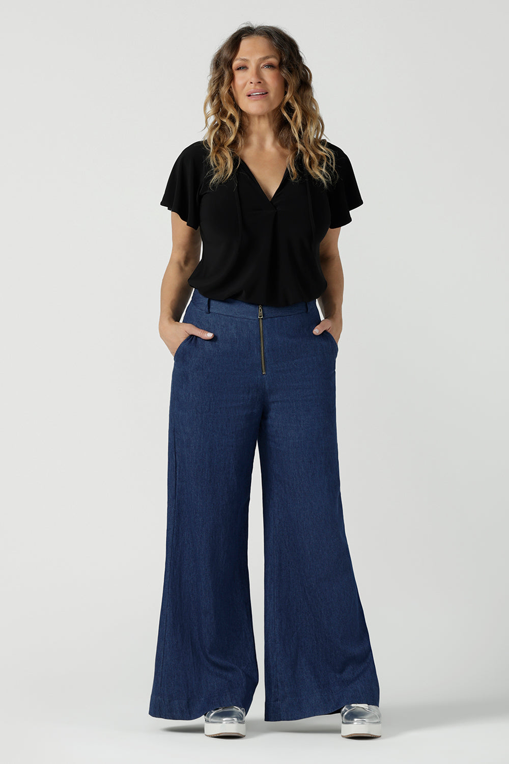 A size 10 woman wears the Bowie Top in Black. A jersey flutter sleeve style with a v-neckline and tie neck. Made in Australia for women 8-24. Styled back with the Dutton Pant in Vintage Denim. 