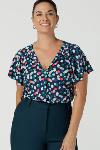 A size 12 happy woman wears a Bowie top with a flutter sleeve  and v-neck with neck ties. All over polka dot print on a soft slinky jersey. Styled back with a Yael Pant in petrol, a high waist tailored work pant. Comfortable work top for women. Made in Australia size 8 - 24.