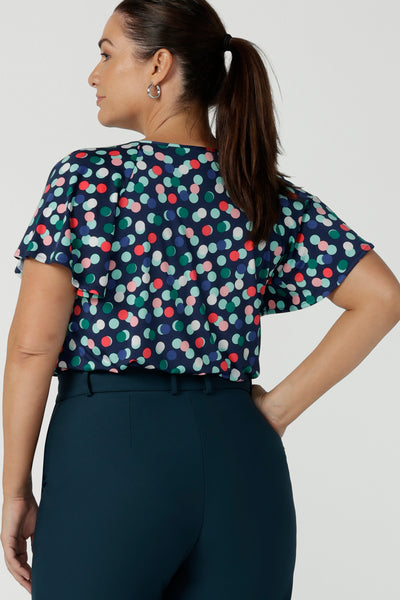 Back view of a size 12 happy woman wears a Bowie top with a flutter sleeve and v-neck with neck ties. All over polka dot print on a soft slinky jersey. Styled back with a Yael Pant in petrol, a high waist tailored work pant. Comfortable work top for women. Made in Australia size 8 - 24.