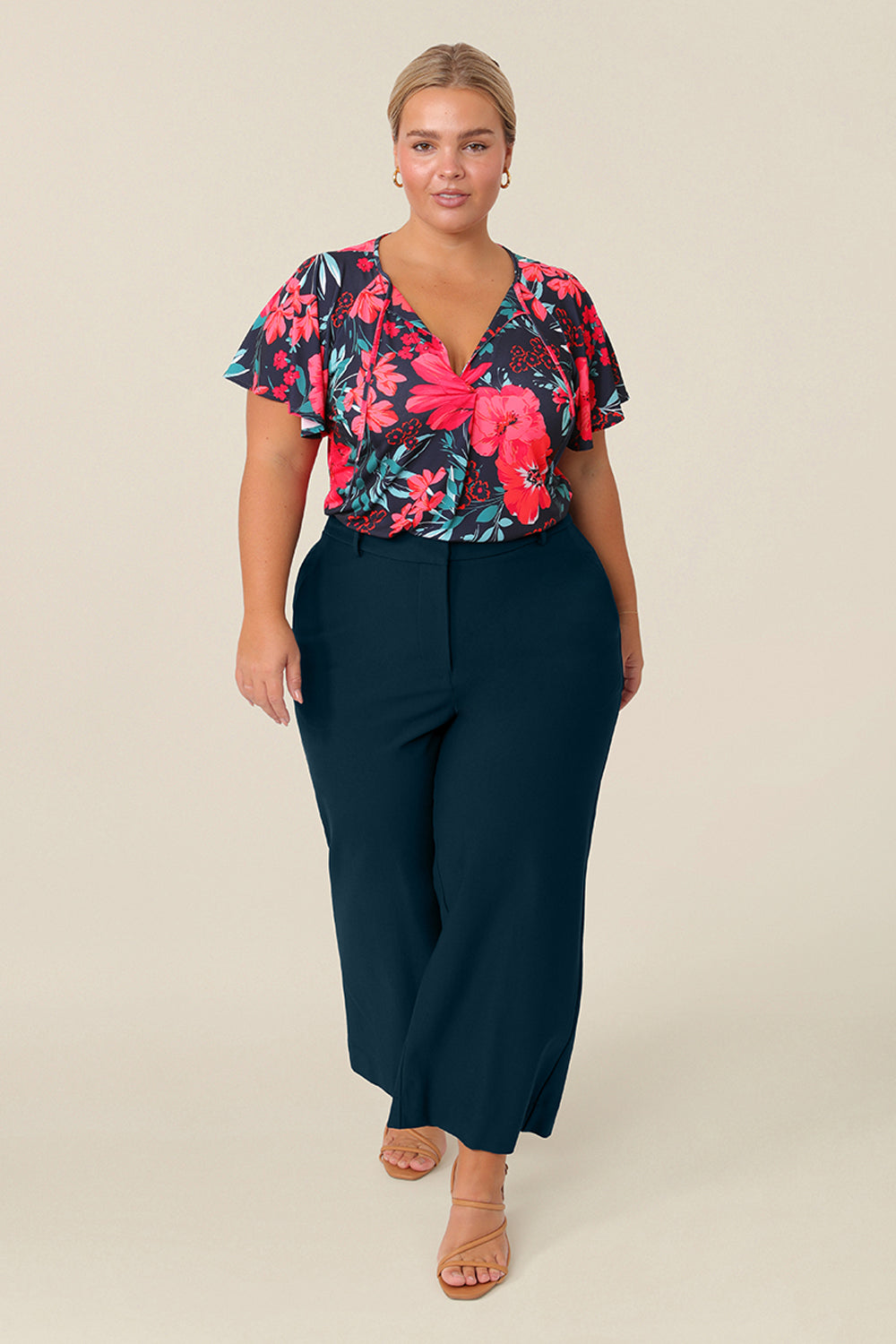 A petite height, size 14 woman wears a casual jersey top. The top has short raglan sleeves, a V-neckline and is printed with a floral pattern paired with petrol wide-leg pants for a casual weekend look.