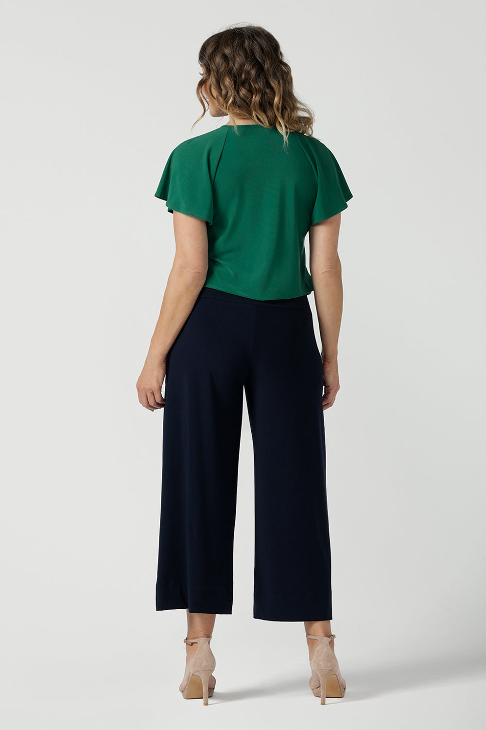 A woman wears the Bowie Top in Jade. A jersey style with flutter sleeve and a v-neckline. Tie neck with a curved hem. A workwear friendly comfortable style for women. Size inclusive and made in Australia for women 8-24. Styled back with black pants.