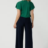 A woman wears the Bowie Top in Jade. A jersey style with flutter sleeve and a v-neckline. Tie neck with a curved hem. A workwear friendly comfortable style for women. Size inclusive and made in Australia for women 8-24. Styled back with black pants.