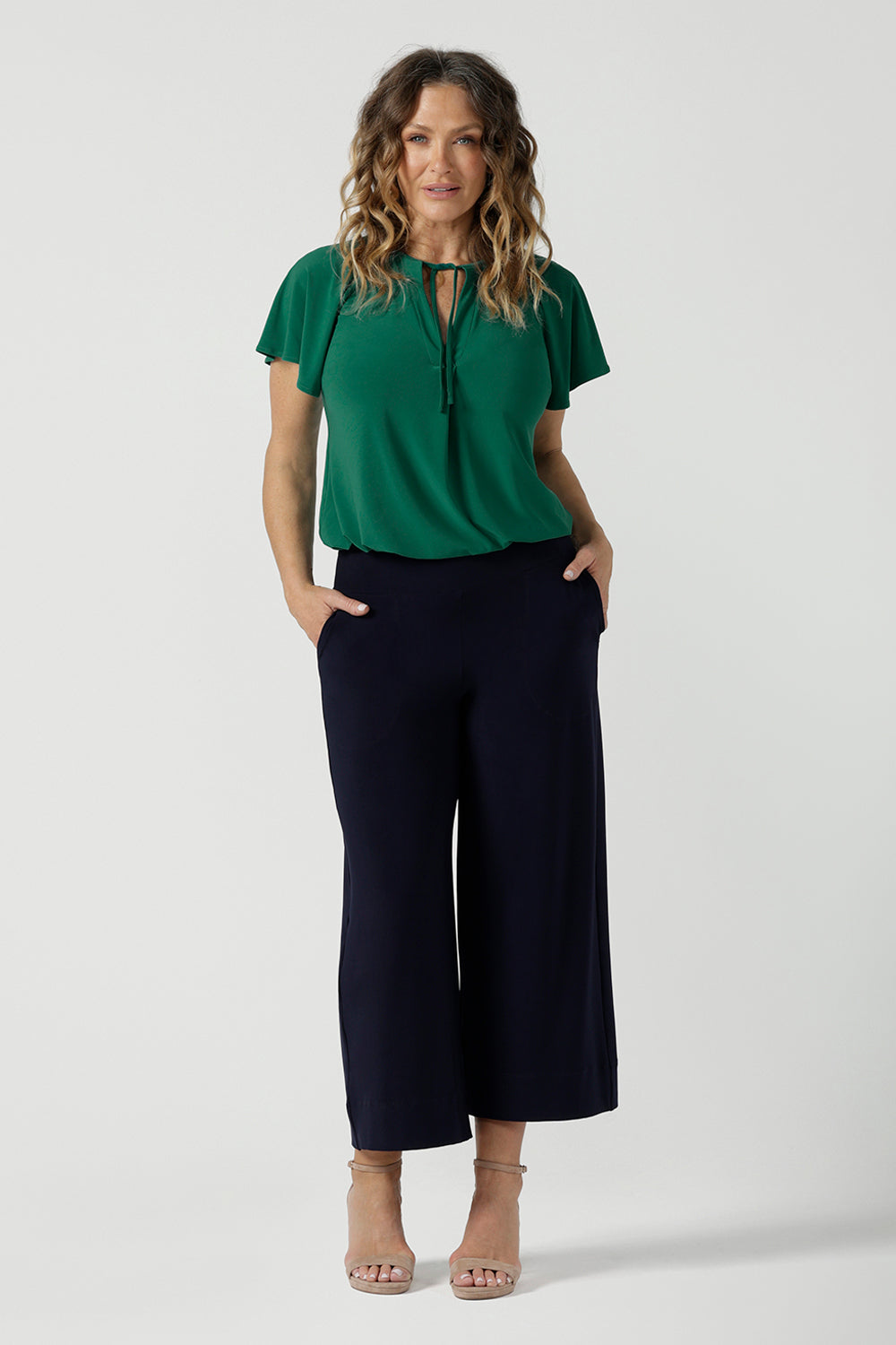 A woman wears the Bowie Top in Jade. A jersey style with flutter sleeve and a v-neckline. Tie neck with a curved hem. A workwear friendly comfortable style for women. Size inclusive and made in Australia for women 8-24. Styled back with black pants.