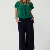 A woman wears the Bowie Top in Jade. A jersey style with flutter sleeve and a v-neckline. Tie neck with a curved hem. A workwear friendly comfortable style for women. Size inclusive and made in Australia for women 8-24. Styled back with black pants.