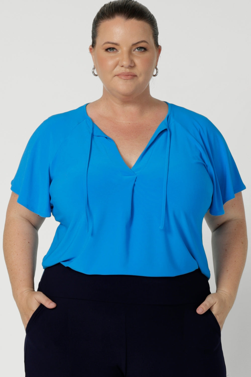 Whether paired with tailored pants or a skirt, it’s a versatile, feel-good piece that’s easy to dress up or down. Feminine style top features v neckline, neckties and short flutter sleeves. Plus size model wears Bowie Top in size 18 paired with navy pants creating an flattering back to work outfit. 