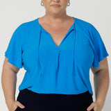 Whether paired with tailored pants or a skirt, it’s a versatile, feel-good piece that’s easy to dress up or down. Feminine style top features v neckline, neckties and short flutter sleeves. Plus size model wears Bowie Top in size 18 paired with navy pants creating an flattering back to work outfit. 