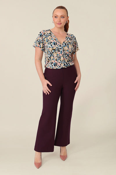 A size 12 curvy woman wears a V-neck short sleeve jersey top in a floral print. She wears the top with flared pants in mulberry. Both are designed and made in Australia for petite to plus size women.