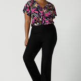 A size 16 woman wears the Bowie top in Vivid Flora with a raglan flutter sleeve, pleat front and v-neckline. Made in Australia for women size 8 - 24. Styled back with Brett pant in navy. 