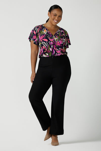 A size 16 woman wears the Bowie top in Vivid Flora with a raglan flutter sleeve, pleat front and v-neckline. Made in Australia for women size 8 - 24. Styled back with Brett pant in navy. 