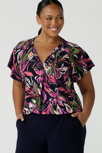 A size 16 woman wears the Bowie top in Vivid Flora with a raglan flutter sleeve, pleat front and v-neckline. Made in Australia for women size 8 - 24.