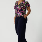 A size 16 woman wears the Bowie top in Vivid Flora with a raglan flutter sleeve, pleat front and v-neckline. Made in Australia for women size 8 - 24. Styled back with Bradley pant in navy. 