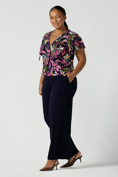 A size 16 woman wears the Bowie top in Vivid Flora with a raglan flutter sleeve, pleat front and v-neckline. Made in Australia for women size 8 - 24. Styled back with Bradley pant in navy. 