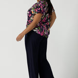 Back view of a size 16 woman wears the Bowie top in Vivid Flora with a raglan flutter sleeve, pleat front and v-neckline. Made in Australia for women size 8 - 24. Styled back with Bradley pant in navy.