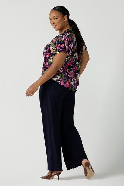 Back view of a size 16 woman wears the Bowie top in Vivid Flora with a raglan flutter sleeve, pleat front and v-neckline. Made in Australia for women size 8 - 24. Styled back with Bradley pant in navy.