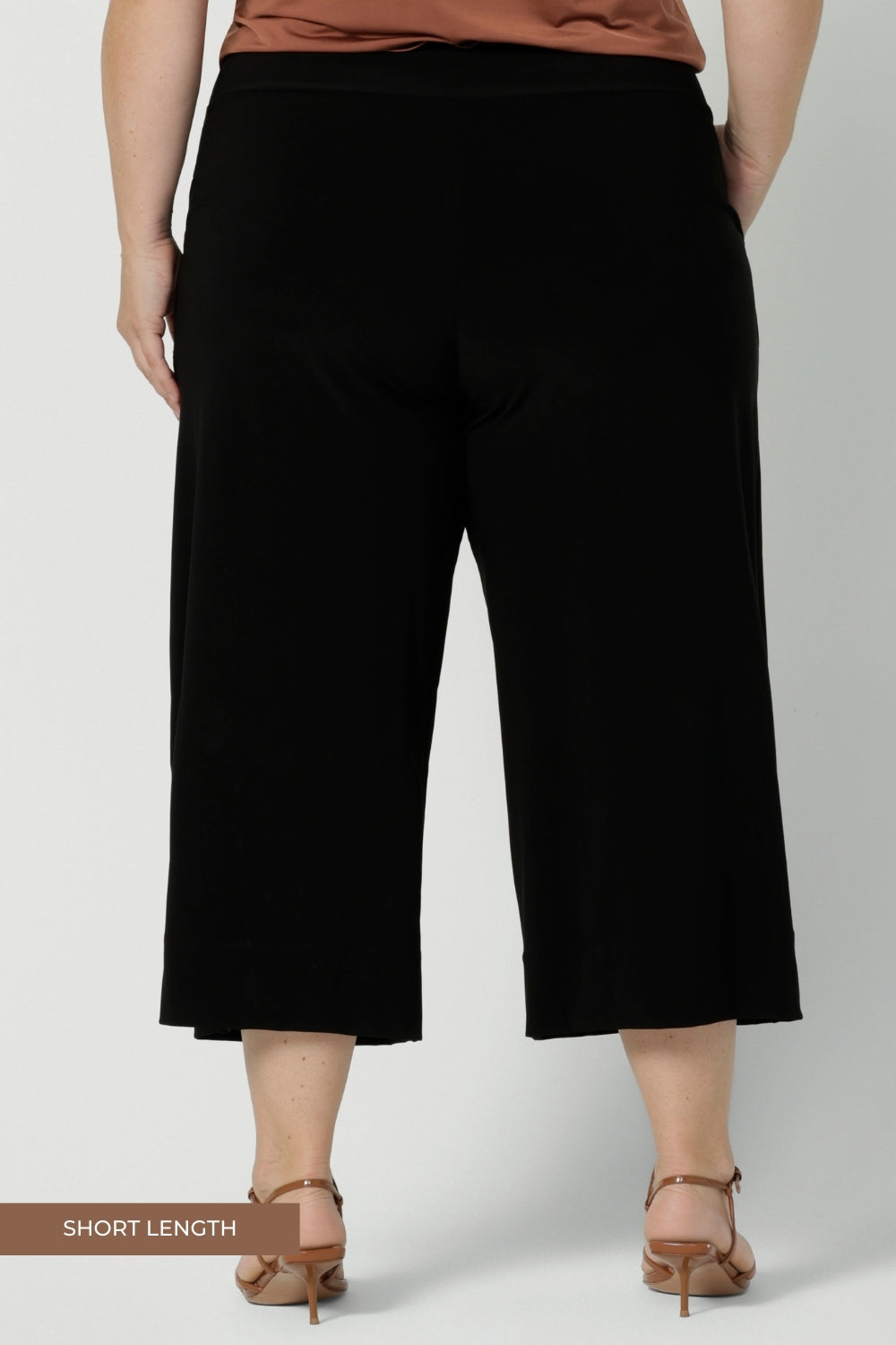 Back view of comfortable, wide leg black culotte pants are worn by a plus size, size 18 woman. Shorter cropped pants with pockets, these pull-on trousers are made in Australia by women's clothing brand, Leina & Fleur in sizes 8-24.