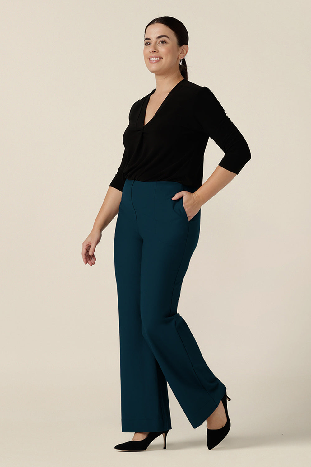 A size 10 woman wears petrol blue, tailored bootcut pants with a long sleeve black jersey top. Made in Australia in ponte fabric, these comfortable work pants are great for corporate women - shop in sizes 8 to 24.