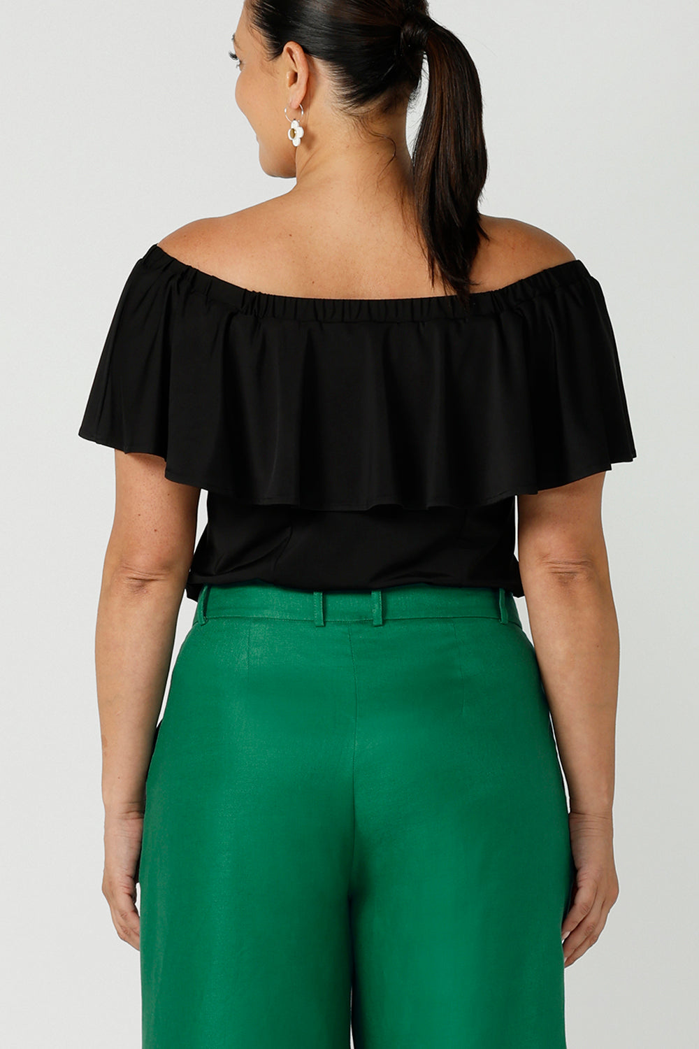 Back view size 12 woman wears an off shoulder ruffle Briar top in black. A versatile top that can be worn off or on shoulder 3 ways. Worn with emerald green linen tailore pants. Great for summer. Designed and made in Australia sizes 8-24.