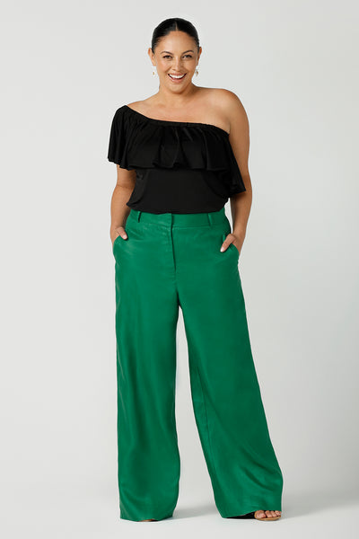 A size 12 woman wears an off shoulder ruffle Briar top in black. A versatile top that can be worn off or on shoulder 3 ways. Worn with emerald green linen tailore pants. Great for summer. Designed and made in Australia sizes 8-24.