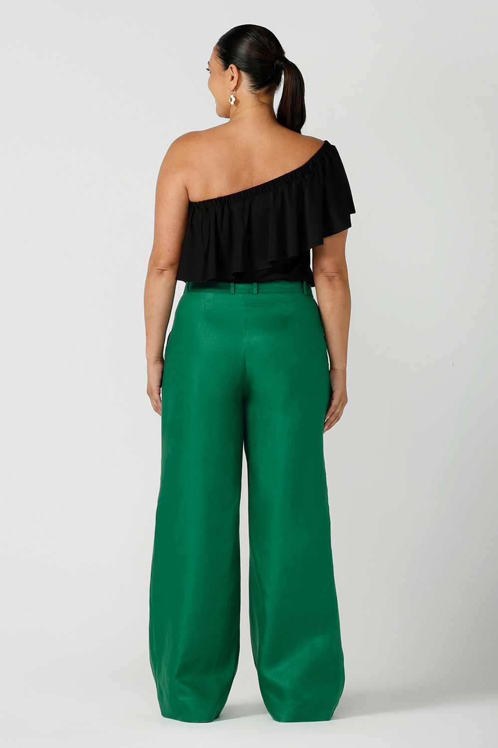 Back view of Back view size 12 woman wears an off shoulder ruffle Briar top in black. A versatile top that can be worn off or on shoulder 3 ways. Worn with emerald green linen tailore pants. Great for summer. Designed and made in Australia sizes 8-24.