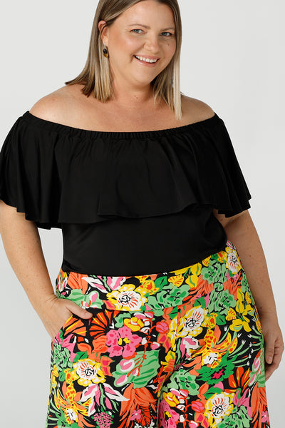 A size 18 woman wears an off shoulder ruffle Briar top in black. A versatile top that can be worn off or on shoulder 3 ways. Worn back with bright and colour Caspian Pant in a soft jersey Cancun print. Great for summer. Designed and made in Australia sizes 8-24.