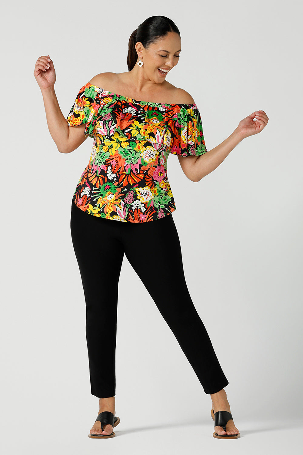 Happy woman wears a size 12 off shoulder ruffle Briar top in the Cancun print.  A bright and colourful pop of green, pink, orange and yellow floral print. Styled back with black work pants. A versatile top for the summer season, wear this top 3 ways. Designed and made in Australia sizes 8-24.