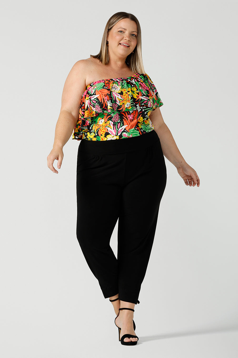 Curvy woman wears a size 18 off shoulder ruffle Briar top in the Cancun print. A bright and colourful pop of green, pink, orange and yellow floral print. Styled back with black work pants. A versatile top for the summer season, wear this top 3 ways. Designed and made in Australia sizes 8-24.