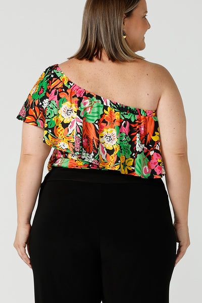 Back view of a curvy woman wears a size 18 off shoulder ruffle Briar top in the Cancun print. A bright and colourful pop of green, pink, orange and yellow floral print. Styled back with black work pants. A versatile top for the summer season, wear this top 3 ways. Designed and made in Australia sizes 8-24.