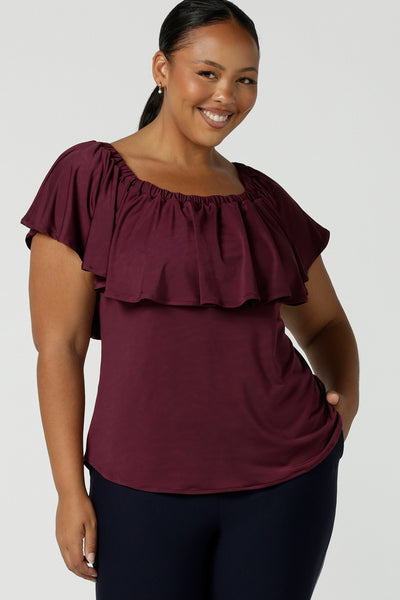 A size 18, plus size woman wears a frill neck, slinky jersey top in plum, with an elasticated neckline worn on the shoulders. Made in Australia by women's clothing brand, Leina and Fleur, this jersey summer top for women is available to shop online in sizes 8 to 24. 