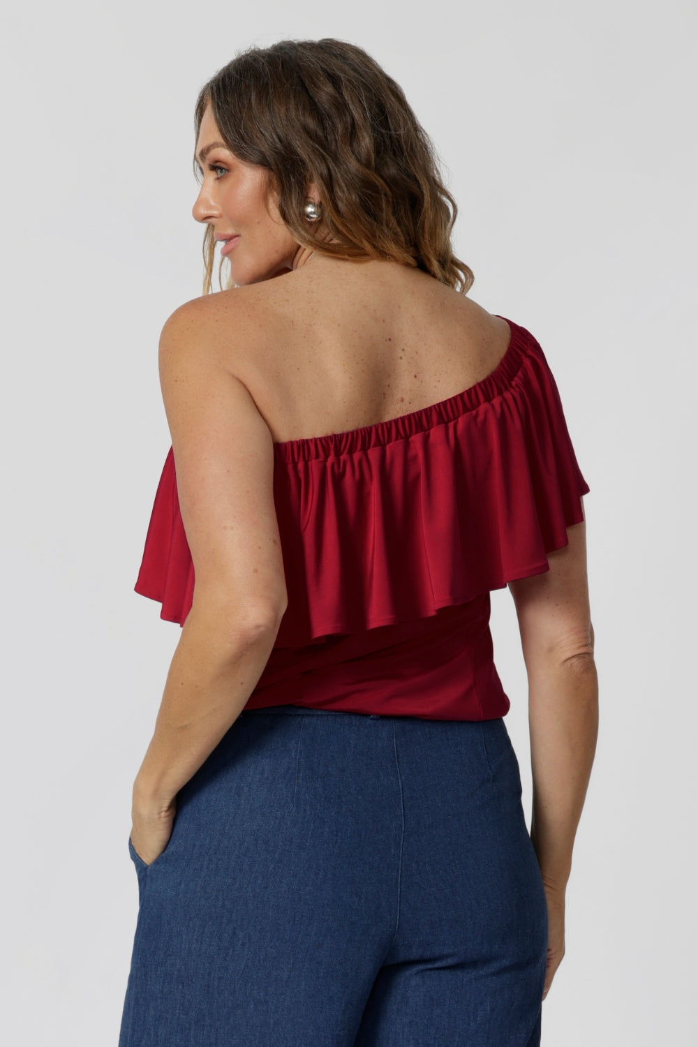 Back view of 40 year old model wears red off the shoulder top. Top has been styled under the shoulders paired back with our Vintage Denim Dutton Pants. Made to drape beautifully with red slinky jersey fabric. The red colour makes it perfect for any season, adding a bold pop of colour. Australian made quality, this easy care top allows you to express your unique style. Retail label Leina and Fleur proudly stocks plus and petite sizes ranging from 8 to 24.