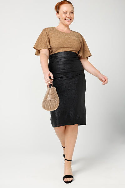 A size 12, fuller figure woman wears a sparkly honey glam top with flutter sleeves. Paired with a shimmery tube skirt for an evening look. Made in Australia by Australian and New Zealand women's clothing company, this shimmer jersey top wears well with evening and occasion-wear skirts, pants and suit jackets.
