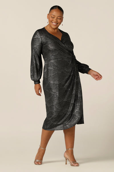 A plus size, size 18 woman wears a metallic cocktail dress for fuller figure women. This long sleeve, wrap dress has a sweetheart neckline and below-the-knee skirt. Made in Australia by Australian and New Zealand women's clothing label, L&F, this semi formal dress is available to shop in sizes 8 to 24.