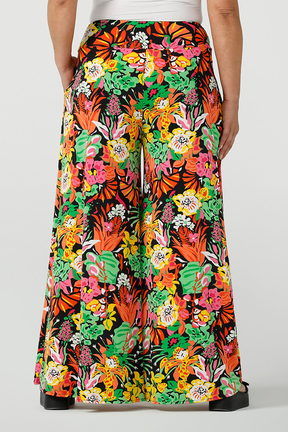 Back view of a size 12 woman wearing the Caspian Palazzo pant n a beautiful bright colourful print. In soft and lightweight jersey this pant features side pocket and a wide leg opening. Styled back with a black cami top and platform sandals. A perfect look for weekend wear. Made in Australia size 8-24.