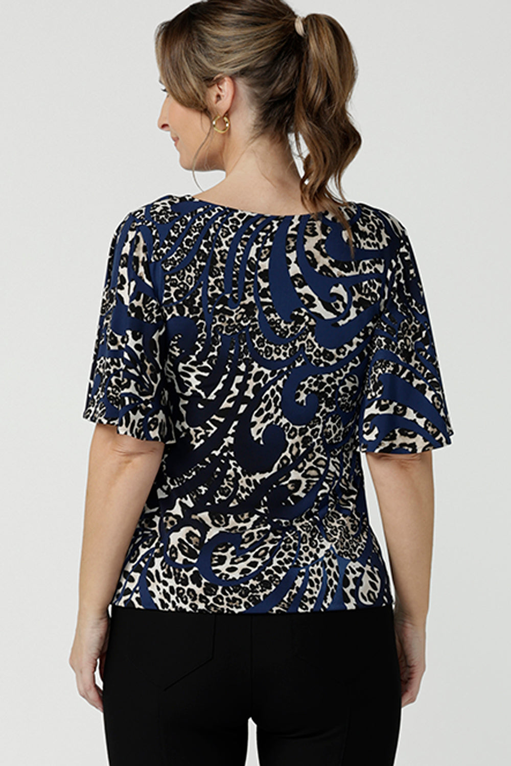 Back view of A size 10 over 40 year old woman wears a jersey top in animal print on a navy base, styled with black, fitted pants. This Australian-made women's top has flutter sleeves, a high, round neckline and and high-low hem perfect for weekend casual and travel capsule wardrobes.