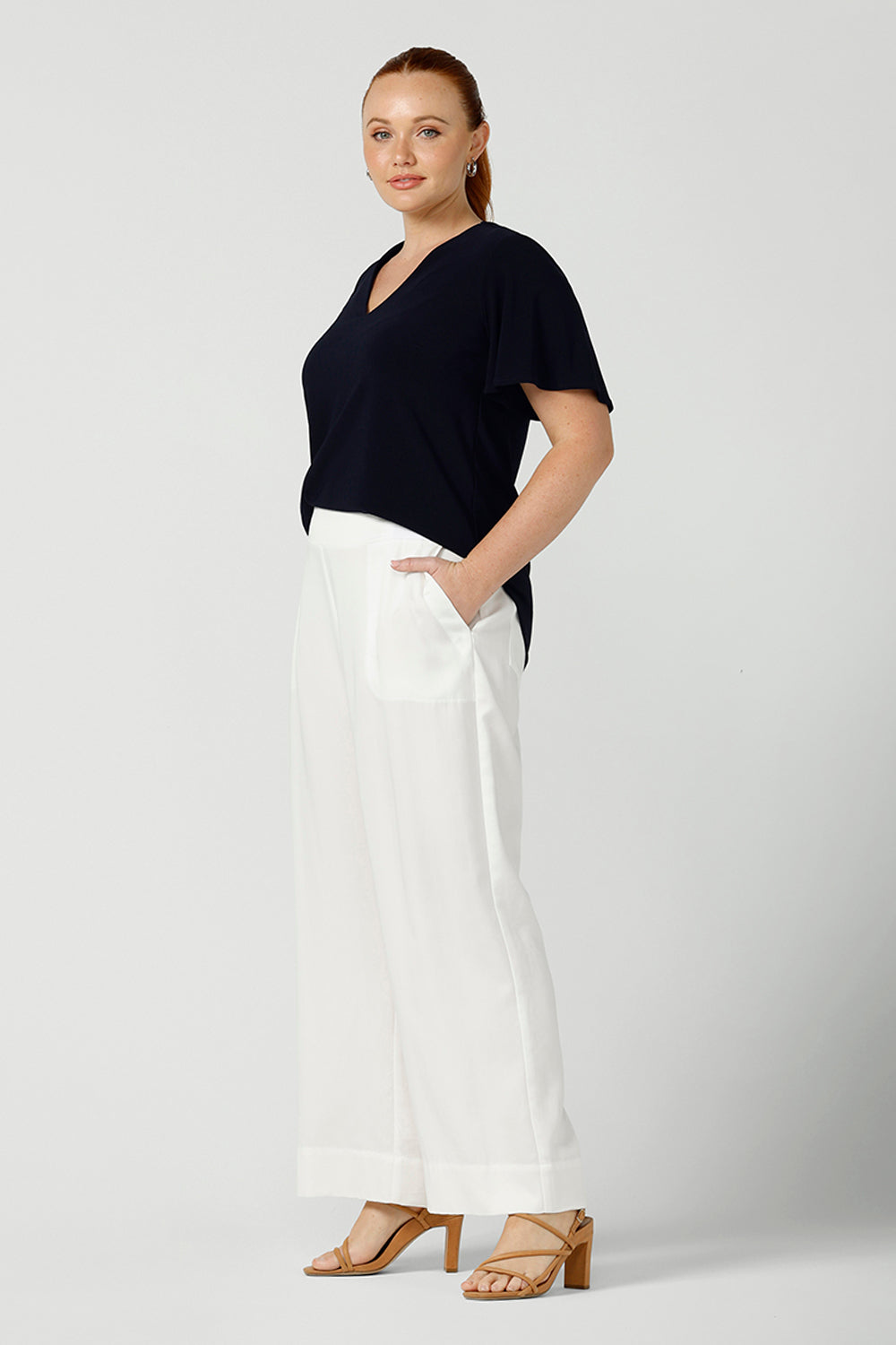 Worn by a size 12, curvy woman, this is a semi-fitted, V-neck top with flutter sleeves in navy blue jersey. Made in Australia by women's clothing brand, Leina & Fleur , this classic navy top is worn with wide leg white pants for summer.  Great for capsule wardrobes, smart casual wear and as a work blouse, shop this top and other women's clothing online in sizes 8 to 24 at L&F's online fashion boutique!