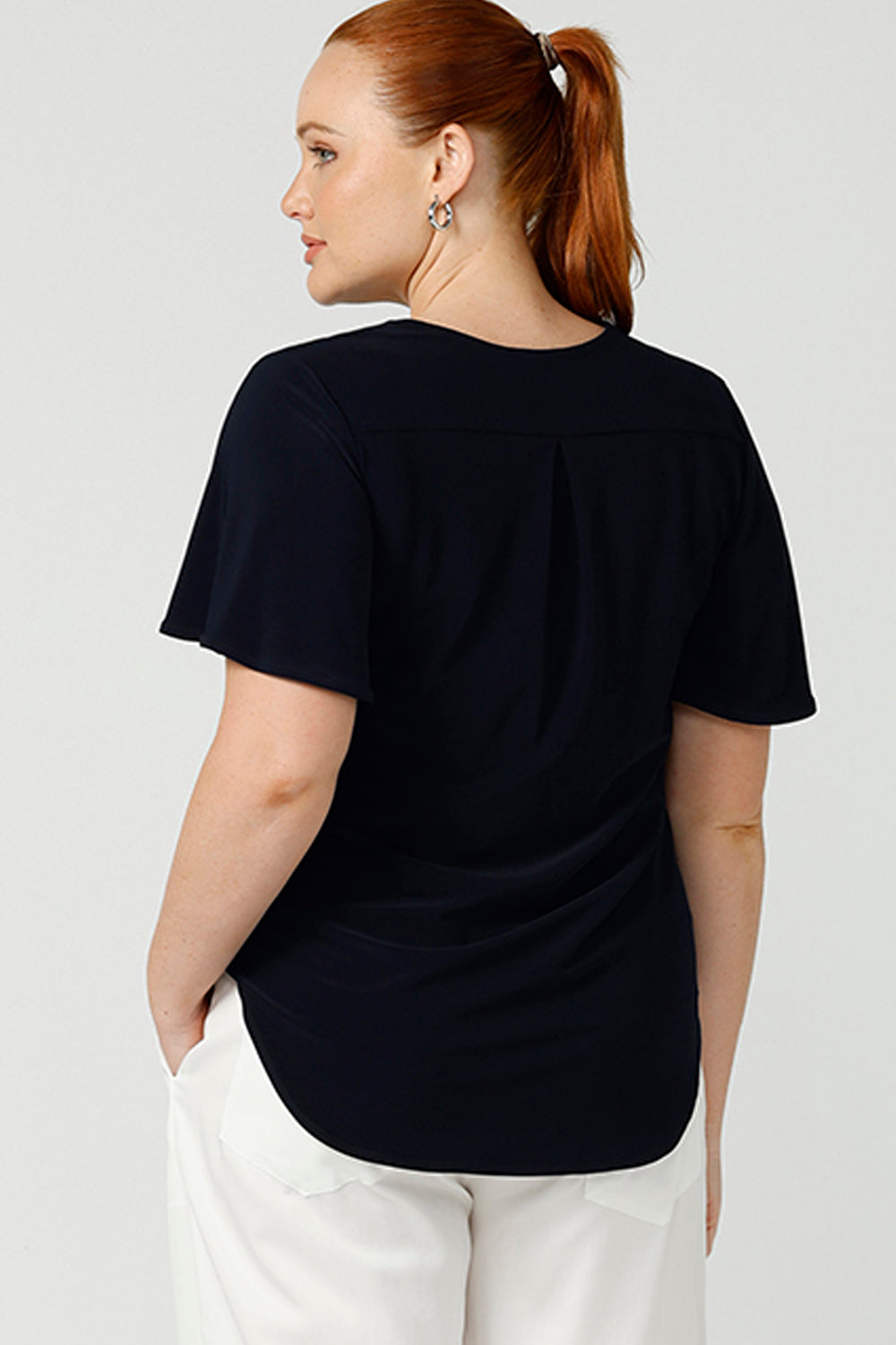 Back view of the shoulder yoke and back pleat of a navy blue jersey top. Worn by a size 12, curvy woman this semi-fitted, V-neck top has flutter sleeves. Made in Australia by women's clothing brand, Leina & Fleur , this classic top is great for capsule wardrobes, smart casual wear and as a work blouse. Shop women's clothing online in sizes 8 to 24 at L&F's online fashion boutique!