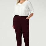 Designed with a mid-rise fit, they feature a waistband with belt loops for added styling options. The invisible front zip with an internal button ensures a sleek, clean look while providing a secure fit. Size 18 woman wears her Corbin Pants with white blouse and brown heels.