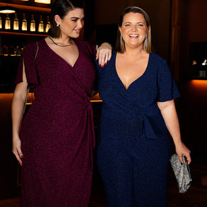 Showing two women dressed in Australian-made fashion for a Christmas party. Woman 1 is a plus size, curvy size 18 woman wearing a raspberry pink cocktail dress with wrap front, tulip skirt and flutter sleeves. Woman 2 is a size 18 woman wearing a plus size, evening wear jumpsuit in sapphire blue lurex fabric with flutter sleeves. Both cocktail dress outfits are available to shop online in inclusive 8 to 24 size range.  