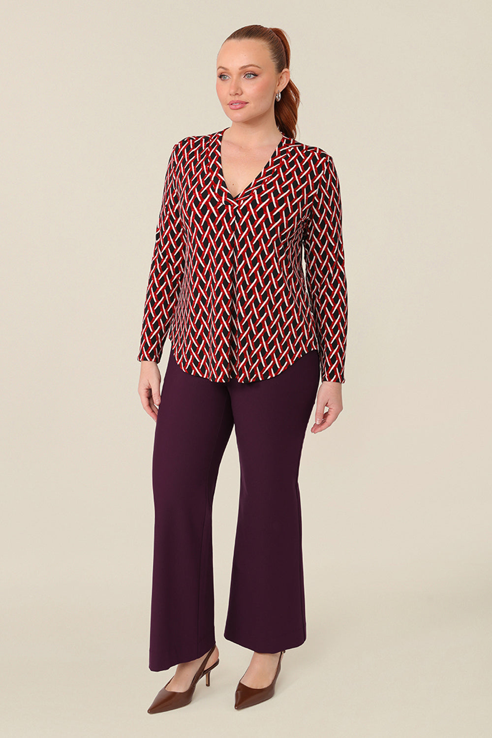 A long sleeve, women's work top, this red, white and black chevron print V-neck top has a shirttail hem. Worn by a curvy woman, this workwear top is worn with mulberry red, flared leg tailored work pants. Shop this office outfit at Australian fashion brand, L&F in petite to plus sizes.