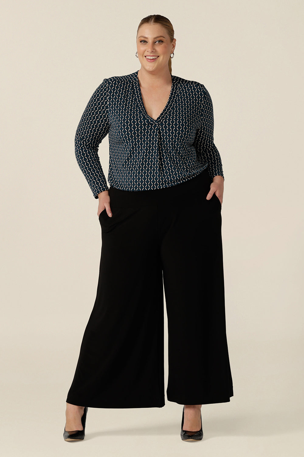 A plus size woman wears black wide leg pants with pockets and a V-neck, long sleeve work top to wear at the office. These pull-on, easy care pants are comfortable for your everyday workwear capsule wardrobe. Shop these black pants online in sizes 8 to 24, petite to plus sizes. 