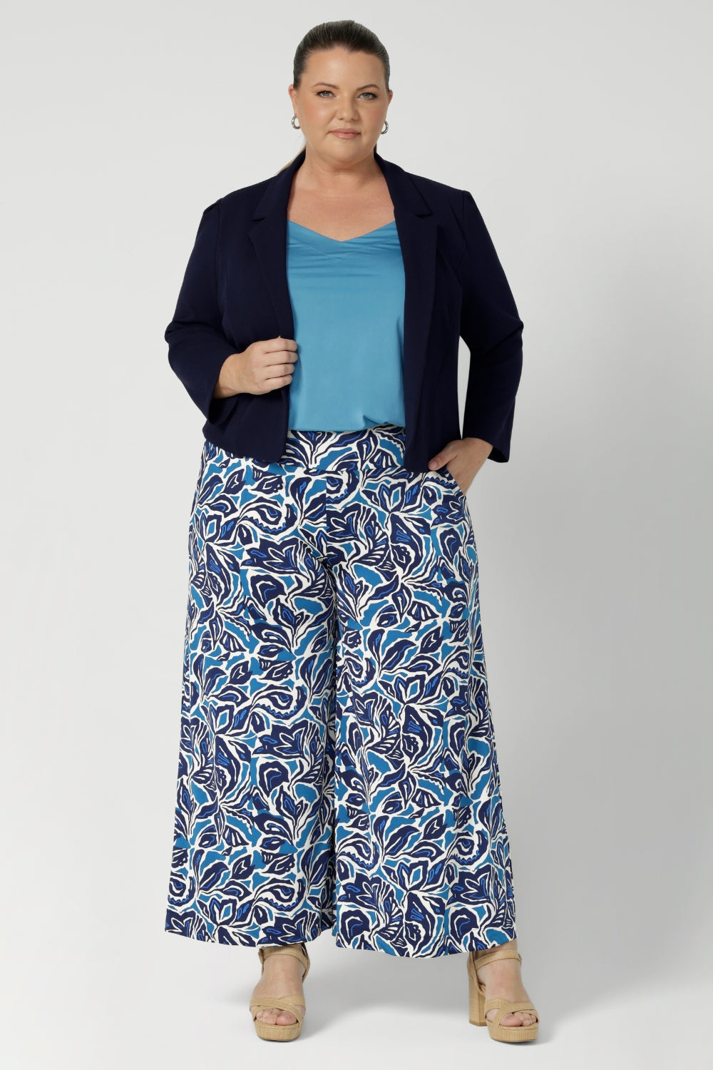 Plus size woman wears wide leg work pants. Danny Culottes offer a sophisticated and comfortable appeal.  Whether you're dressing up for work or embracing a laid-back weekend look, these pants bring a touch of elegance to any occasion. Model has styled her wide leg, printed pants with matching blue cami and navy cropped jacket with a neutral heel & silver jewellery.