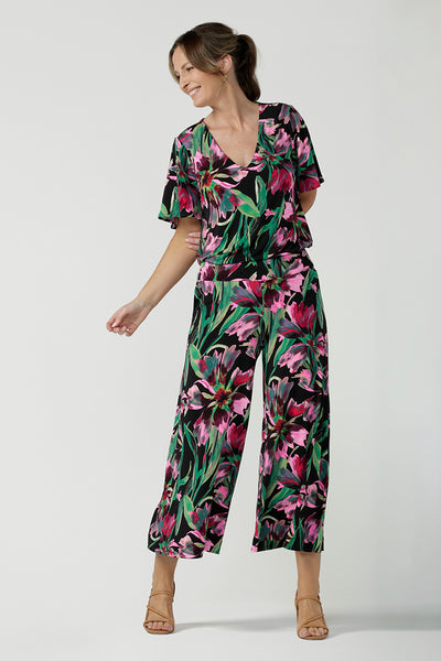 A size 10, 40 plus woman wears a floral print jersey, V-neck top with flutter sleeves as a jumpsuit with wide leg, floral print pants. A good top for summer casual wear, or style tucked as a workwear top. Shop made in Australia tops in petite to plus sizes online at Australian fashion brand, Leina & Fleur.