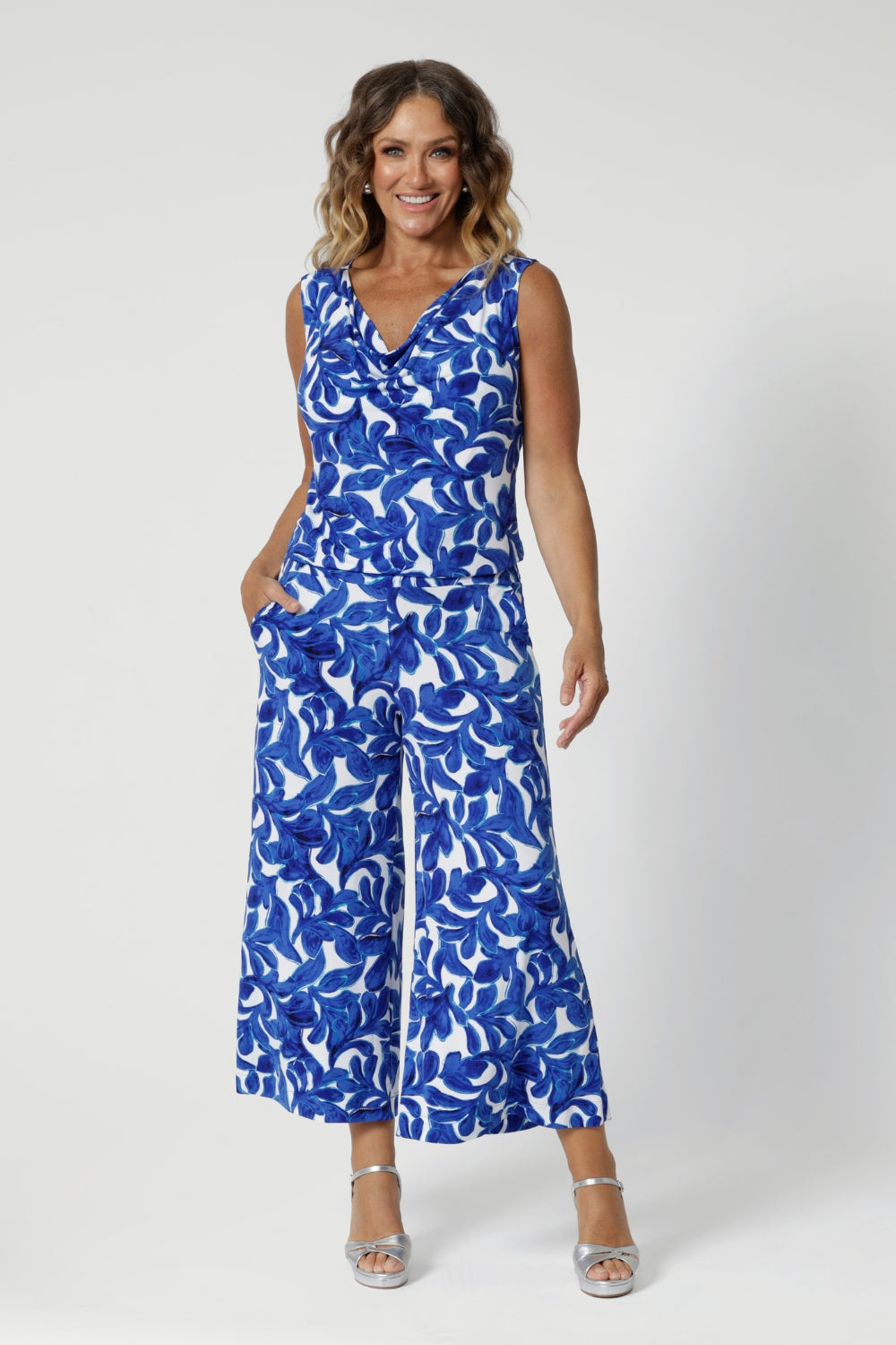 Size 12 woman wears wide leg printed pants made with lightweight dry-touch jersey with print details in shades of blue leaves. Summer ready pants have been styles with matching sleeveless top for a faux jumpsuit look, paired with tall silver heels for an elevated look for summer outings.