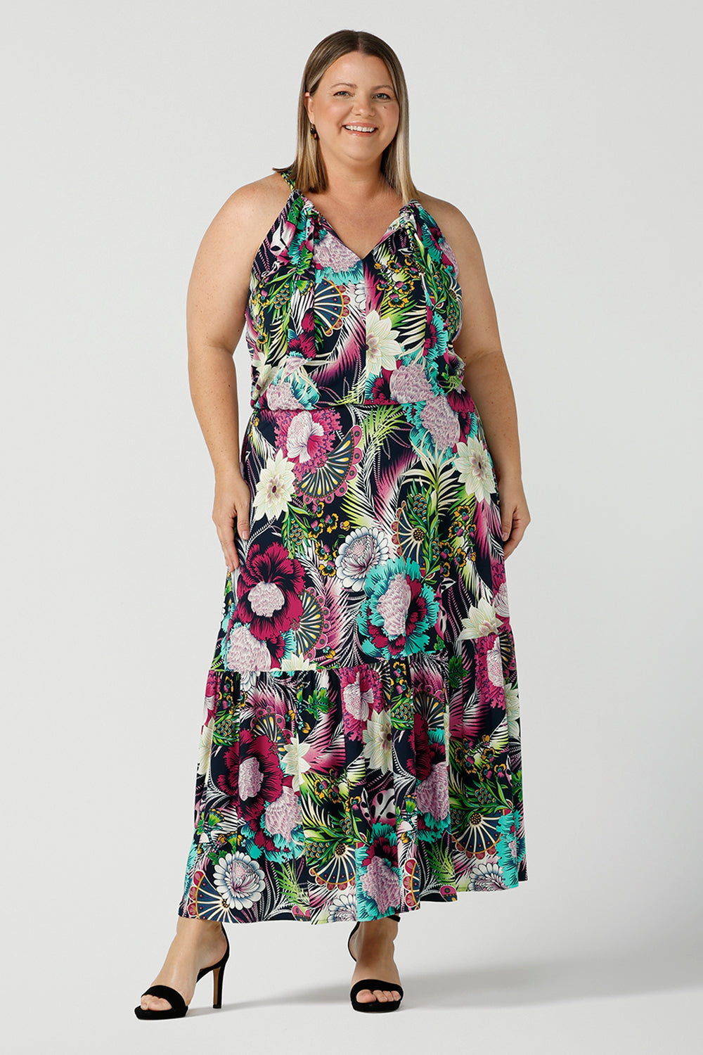 A happy woman wears the Darci dress in Navy Getaway halter neck, jersey maxi dress with ruffle on hem. A detailed tropical print on a navy base with fuchsia, green, yellow and aqua. Pictured on a curvy woman size 18. Made in Australia for women size 8 - 24.