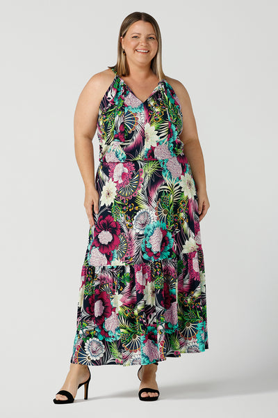 A happy woman wears the Darci dress in Navy Getaway halter neck, jersey maxi dress with ruffle on hem. A detailed tropical print on a navy base with fuchsia, green, yellow and aqua. Pictured on a curvy woman size 18. Made in Australia for women size 8 - 24.