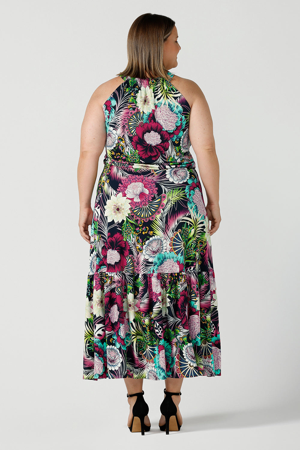 Back view of a woman wearing the Darci dress in Navy Getaway halter neck, jersey maxi dress with ruffle on hem. A detailed tropical print on a navy base with fuchsia, green, yellow and aqua. Pictured on a curvy woman size 18. Made in Australia for women size 8 - 24.