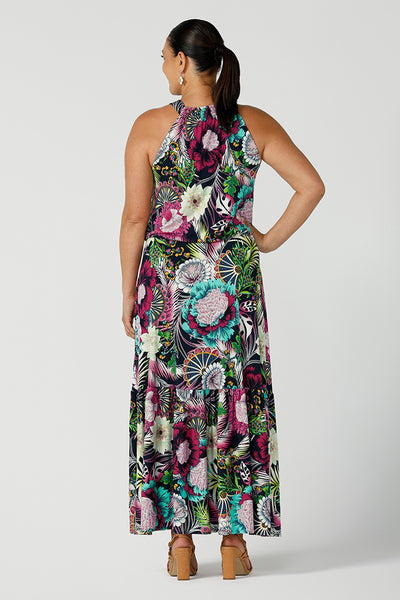 Back view of a woman wearing the Darci dress in Navy Getaway halter neck, jersey maxi dress with ruffle on hem. A detailed tropical print on a navy base with fuchsia, green, yellow and aqua. Pictured on a curvy woman size 12. Made in Australia for women size 8 - 24.