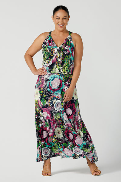 A happy woman wears the Darci dress in Navy Getaway halter neck, jersey maxi dress with ruffle on hem.  A detailed tropical print on a navy base with fuchsia, green, yellow and aqua. Pictured on a curvy woman size 12. Made in Australia for women size 8 - 24.