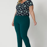 Showing a good summer top for plus size women, a curvy, size 18 woman wears a cowl neck top in aqua blue floral print. A short sleeve top , its worn with tapered leg work pants in green as  a workwear top. This comfortable top is made from stretch jersey and is an easy-care top style. Made in Australia by Australian and New Zealand women's clothes brand, Leina & Fleur.