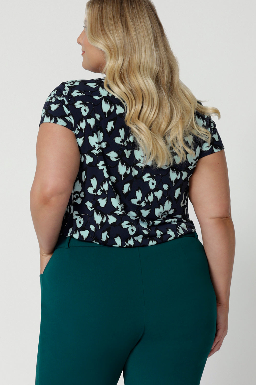 Showing the back view of a good summer top for plus size women, a curvy, size 18 woman wears a cowl neck top in aqua blue floral print. A short sleeve top , its worn with tapered leg work pants in green as  a workwear top. This comfortable top is made from stretch jersey and is an easy-care top style. Made in Australia by Australian and New Zealand women's fashion brand, Leina & Fleur.