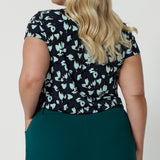 Showing the back view of a good summer top for plus size women, a curvy, size 18 woman wears a cowl neck top in aqua blue floral print. A short sleeve top , its worn with tapered leg work pants in green as  a workwear top. This comfortable top is made from stretch jersey and is an easy-care top style. Made in Australia by Australian and New Zealand women's fashion brand, Leina & Fleur.